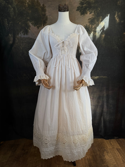 A long ivory/antique-white historical-style chemise dress displayed on a mannequin against a dark painted landscape backdrop. The dress has long sleeves with ruffled cuffs, a smocked bodice, a lace-trimmed neckline with front corset lacing, and lace inset details along the hem.