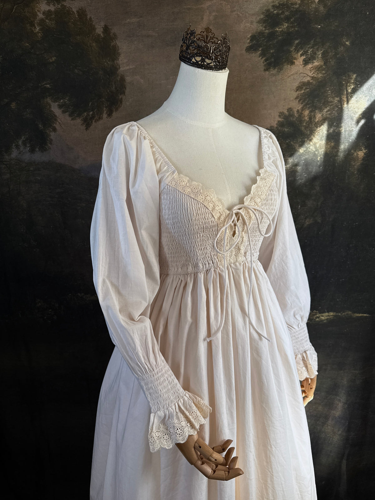 A long ivory/antique-white historical-style chemise dress displayed on a mannequin against a dark painted landscape backdrop. The dress has long sleeves with ruffled cuffs, a smocked bodice, a lace-trimmed neckline with front corset lacing, and lace inset details along the hem.