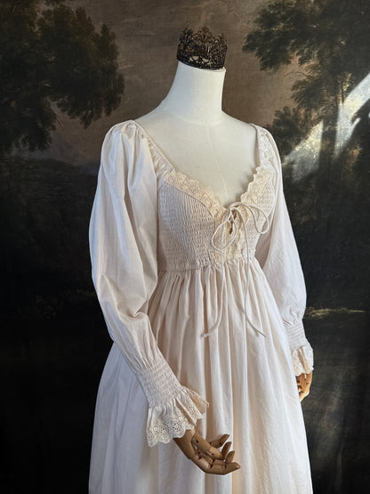 A long ivory/antique-white historical-style chemise dress displayed on a mannequin against a dark painted landscape backdrop. The dress has long sleeves with ruffled cuffs, a smocked bodice, a lace-trimmed neckline with front corset lacing, and lace inset details along the hem.