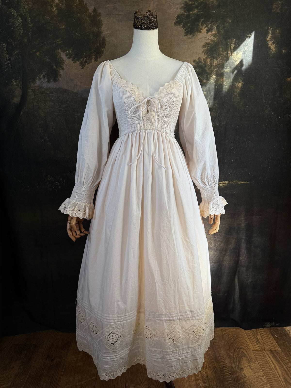 A long ivory/antique-white historical-style chemise dress displayed on a mannequin against a dark painted landscape backdrop. The dress has long sleeves with ruffled cuffs, a smocked bodice, a lace-trimmed neckline with front corset lacing, and lace inset details along the hem.