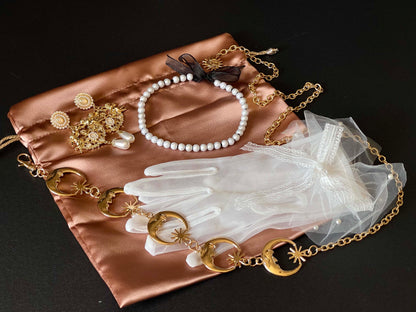 An array of historically inspired accessories - pearl necklace, moon belt, baroque earrings, and chiffon gloves on a satin bag, displaying a possible mystery bag combo.