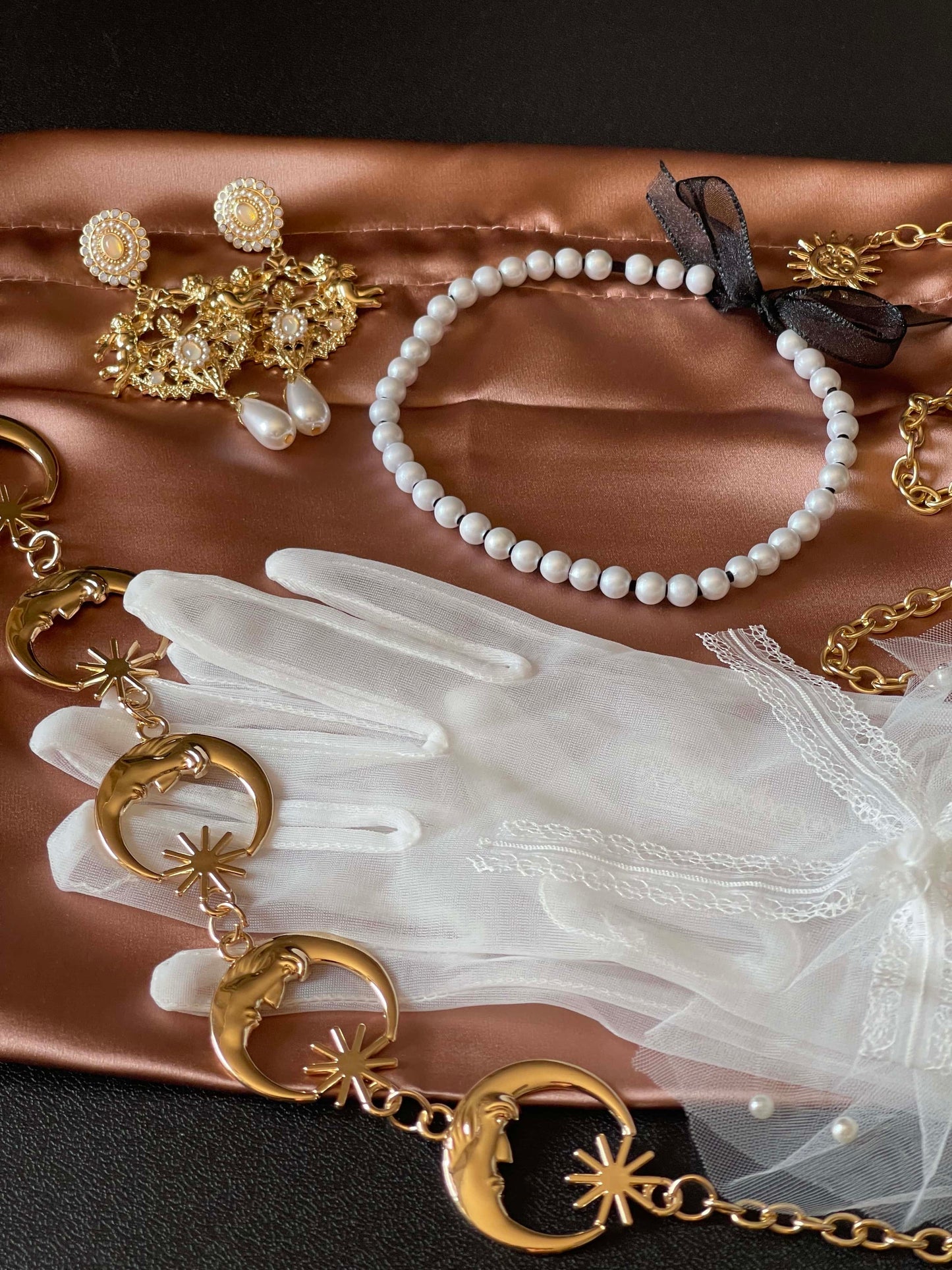 An array of historically inspired accessories - pearl necklace, moon belt, baroque earrings, and chiffon gloves on a satin bag, displaying a possible mystery bag combo.
