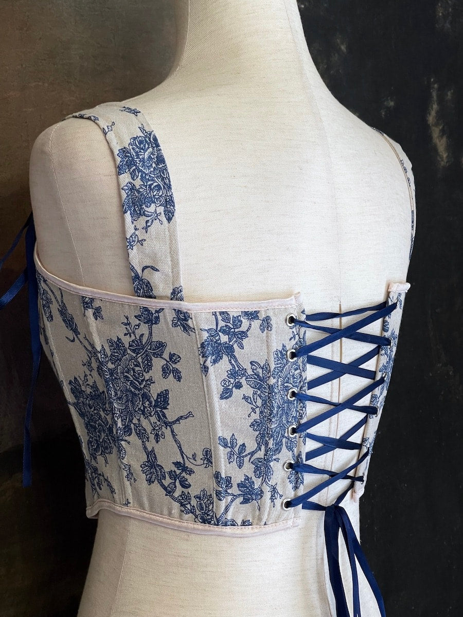 A rococo and regency era inspired blue and ivory colored floral toile de jouy corset pair of stays in front of a historical patterned wallpaper.