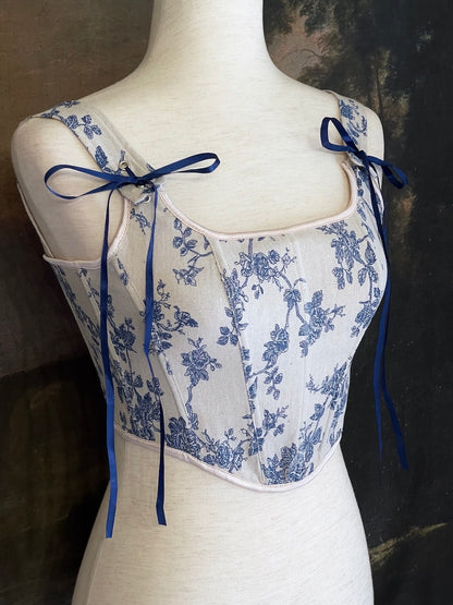 A rococo and regency era inspired blue and ivory colored floral toile de jouy corset pair of stays in front of a historical patterned wallpaper.