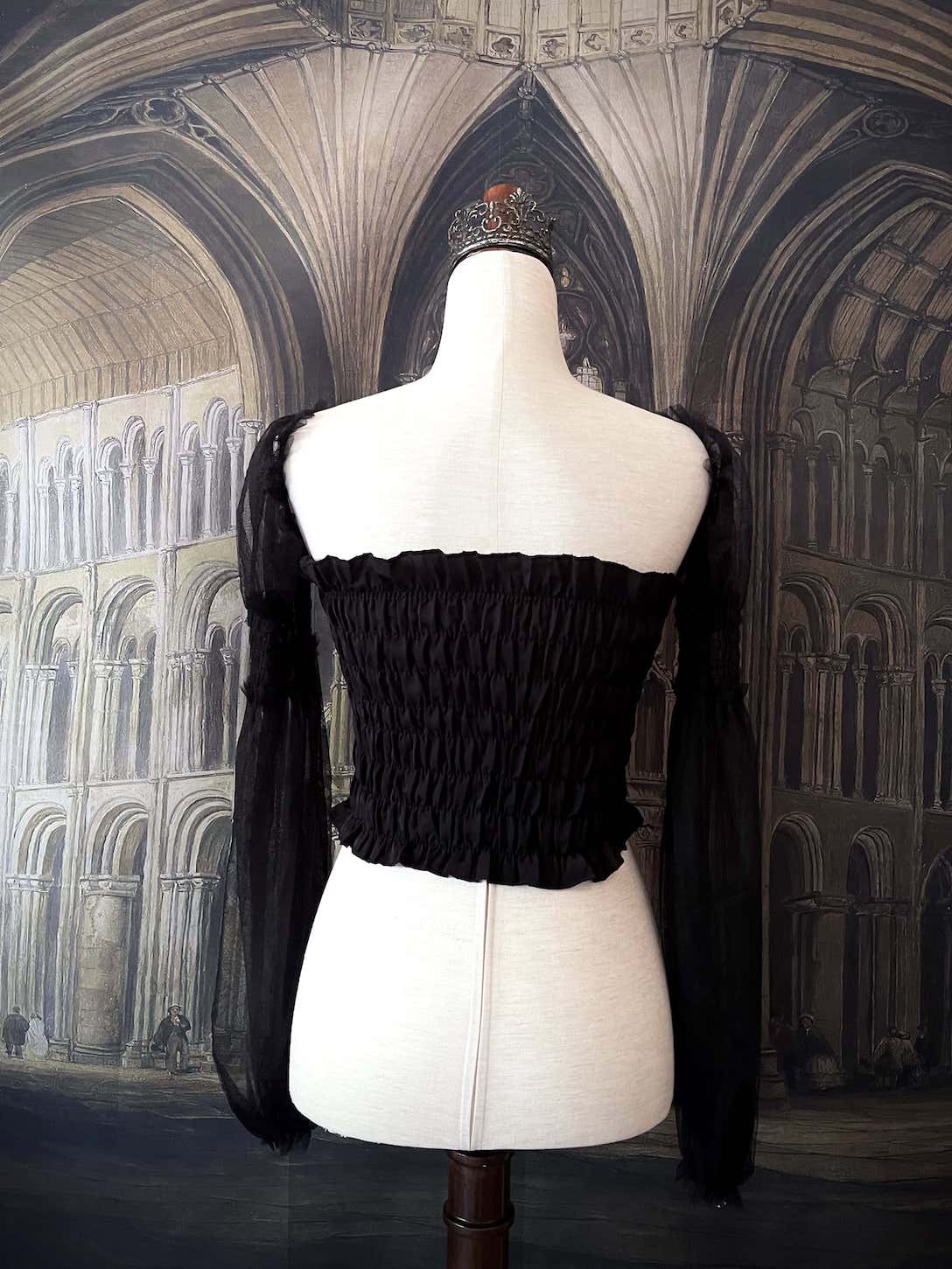 A historically inspired gothic black smocked blouse with mesh juliet sleeves is pictured on a mannequin in front of a medieval gothic cathedral backdrop.