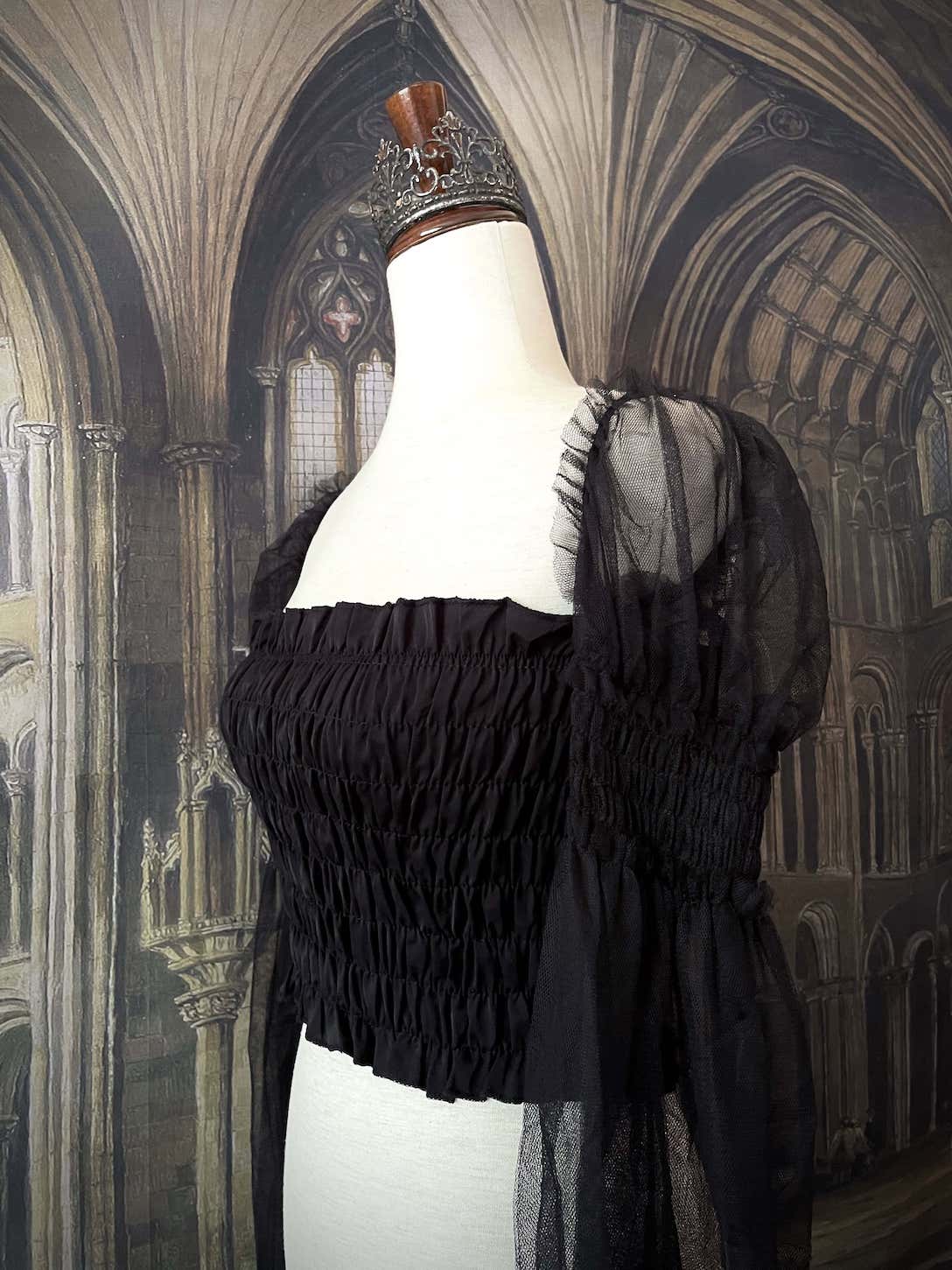 A historically inspired gothic black smocked blouse with mesh juliet sleeves is pictured on a mannequin in front of a medieval gothic cathedral backdrop.