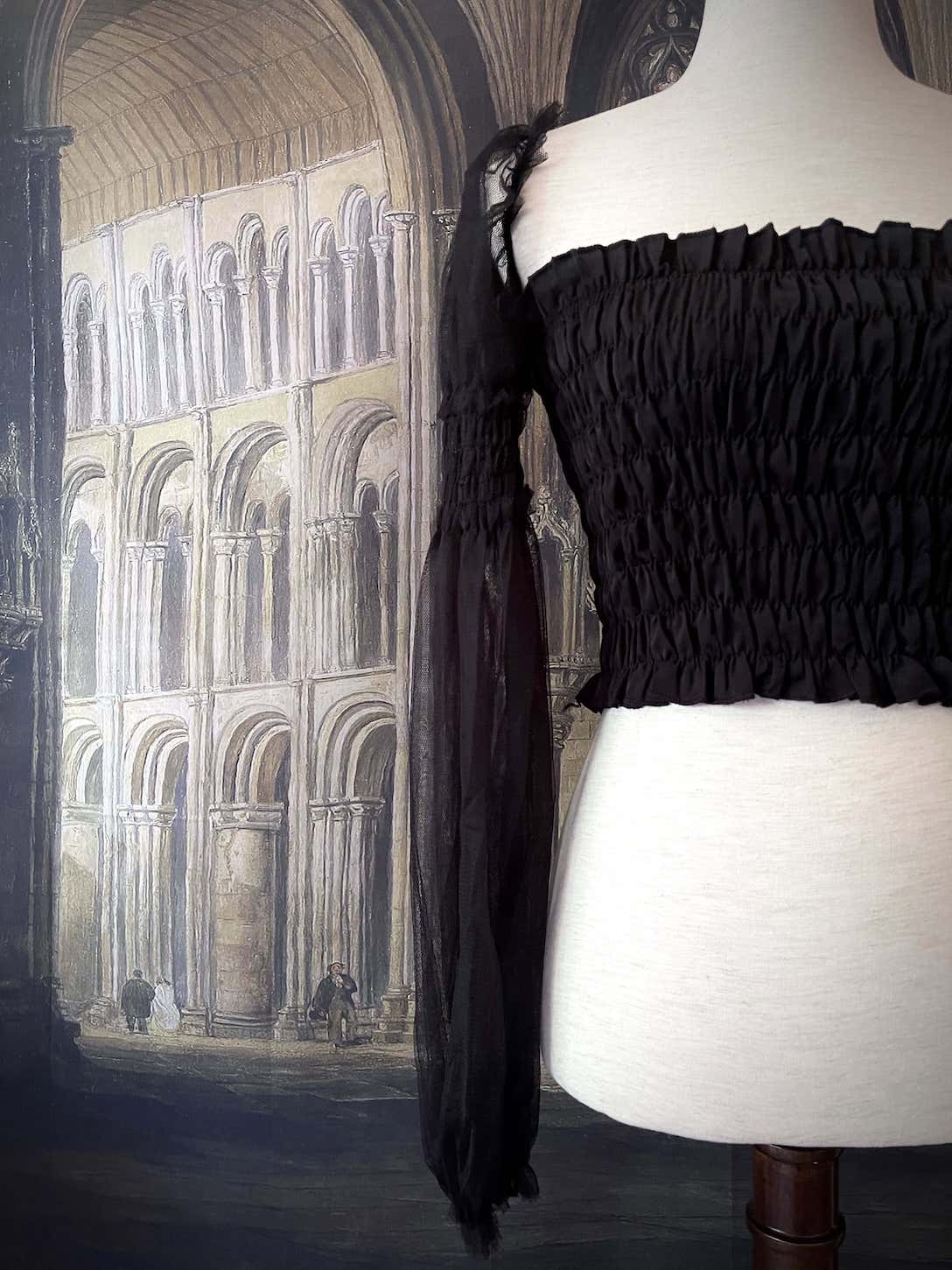 A historically inspired gothic black smocked blouse with mesh juliet sleeves is pictured on a mannequin in front of a medieval gothic cathedral backdrop.