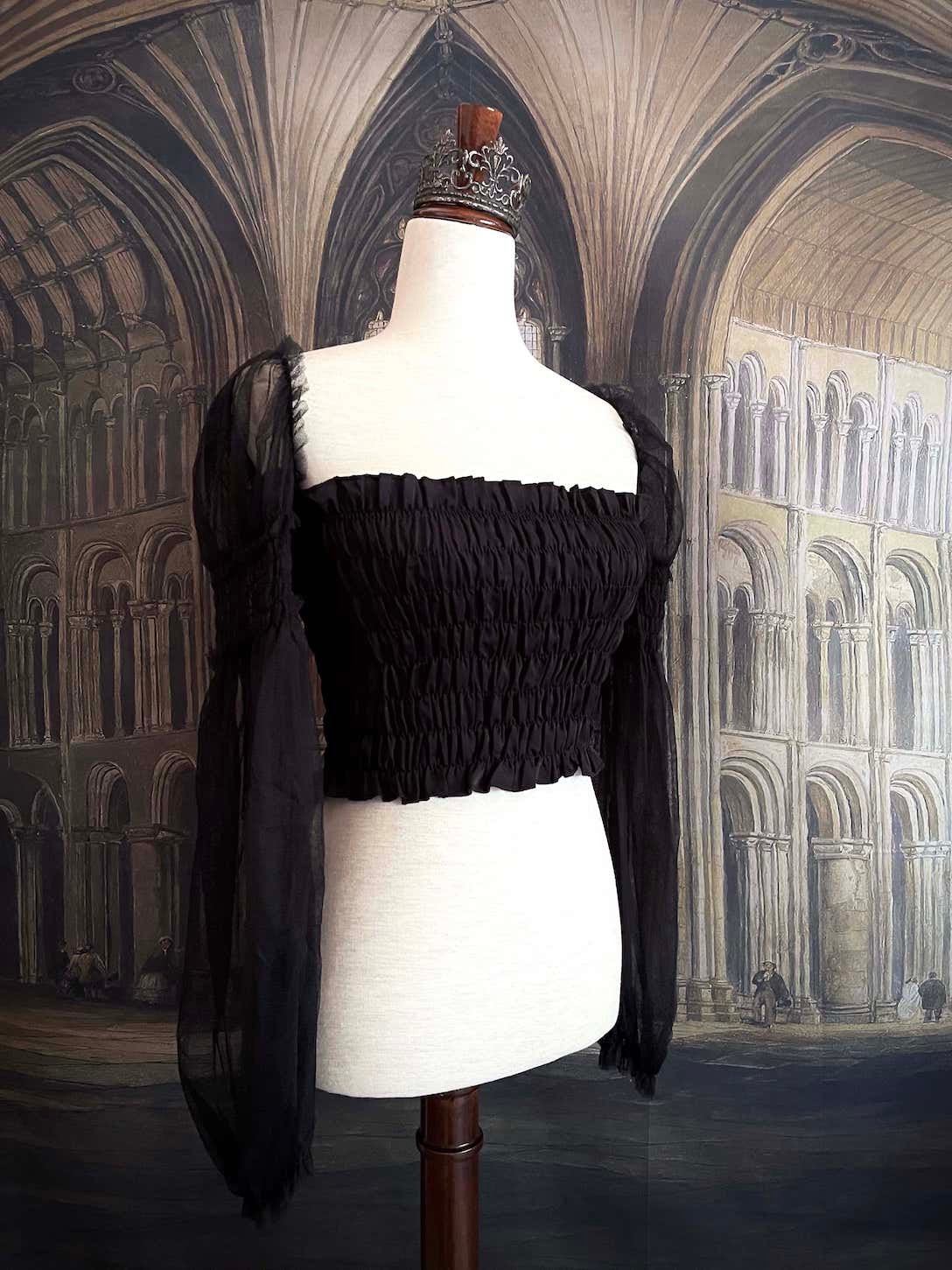 A historically inspired gothic black smocked blouse with mesh juliet sleeves is pictured on a mannequin in front of a medieval gothic cathedral backdrop.