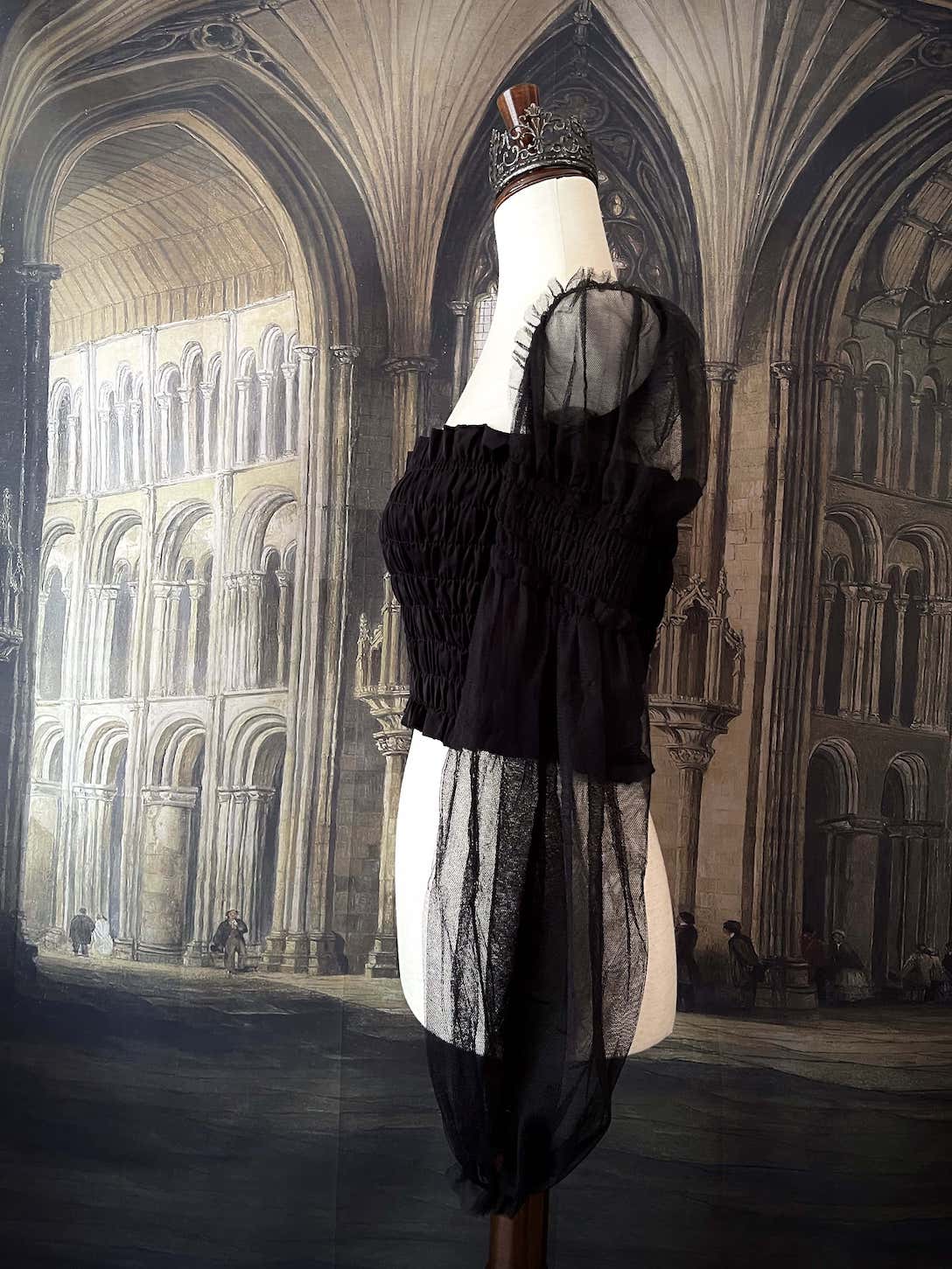 A historically inspired gothic black smocked blouse with mesh juliet sleeves is pictured on a mannequin in front of a medieval gothic cathedral backdrop.