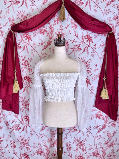 A historically inspired pure white smocked blouse with mesh juliet sleeves is pictured on a mannequin in front of a red and white toile wallpaper.