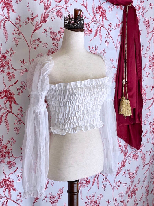 A historically inspired pure white smocked blouse with mesh juliet sleeves is pictured on a mannequin in front of a red and white toile wallpaper.
