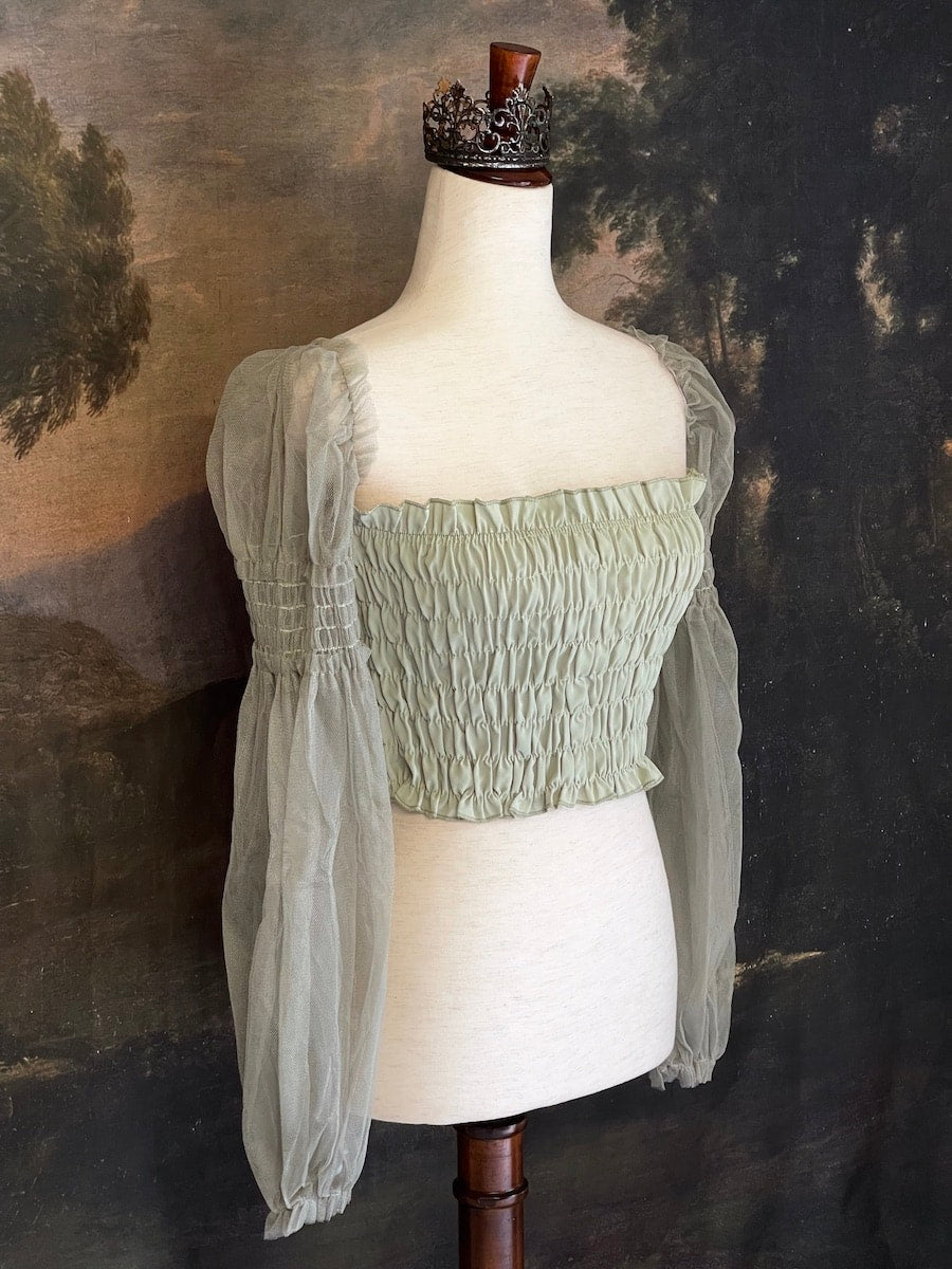 A historically inspired sage green smocked blouse with mesh juliet sleeves is pictured on a mannequin in front of a painted landscape wallpaper.