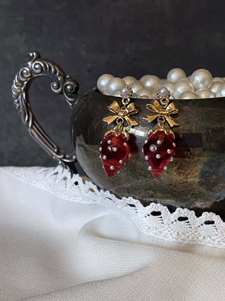 A pair of cottagecore vintage style bow and strawberry earrings in gold metal and red enamel with pearl accents. On an antique silver dish.