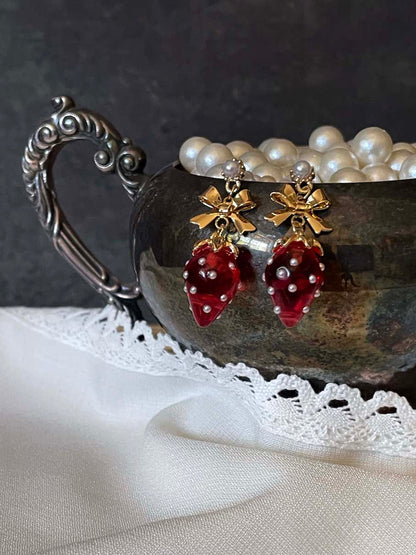 A pair of cottagecore vintage style bow and strawberry earrings in gold metal and red enamel with pearl accents. On an antique silver dish.