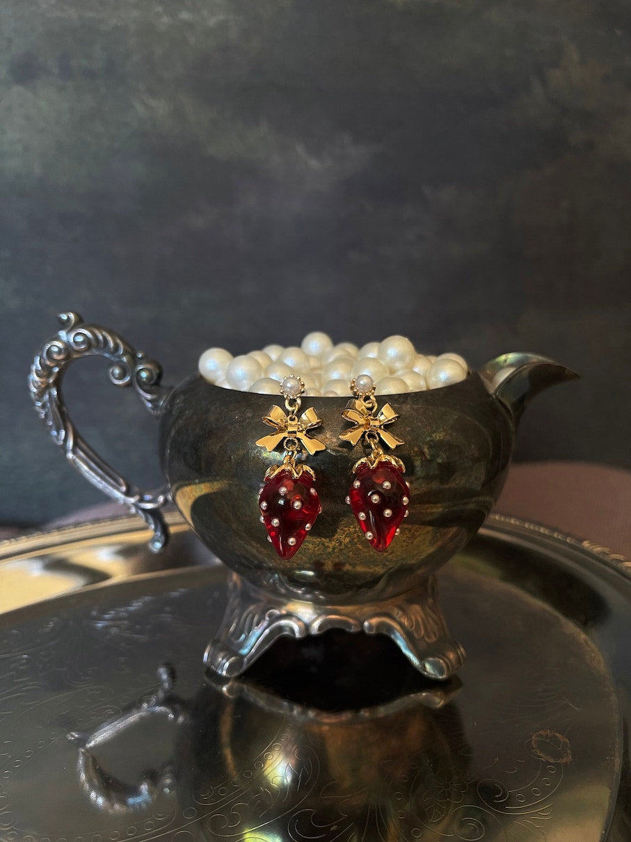 A pair of cottagecore vintage style bow and strawberry earrings in gold metal and red enamel with pearl accents. On an antique silver dish.