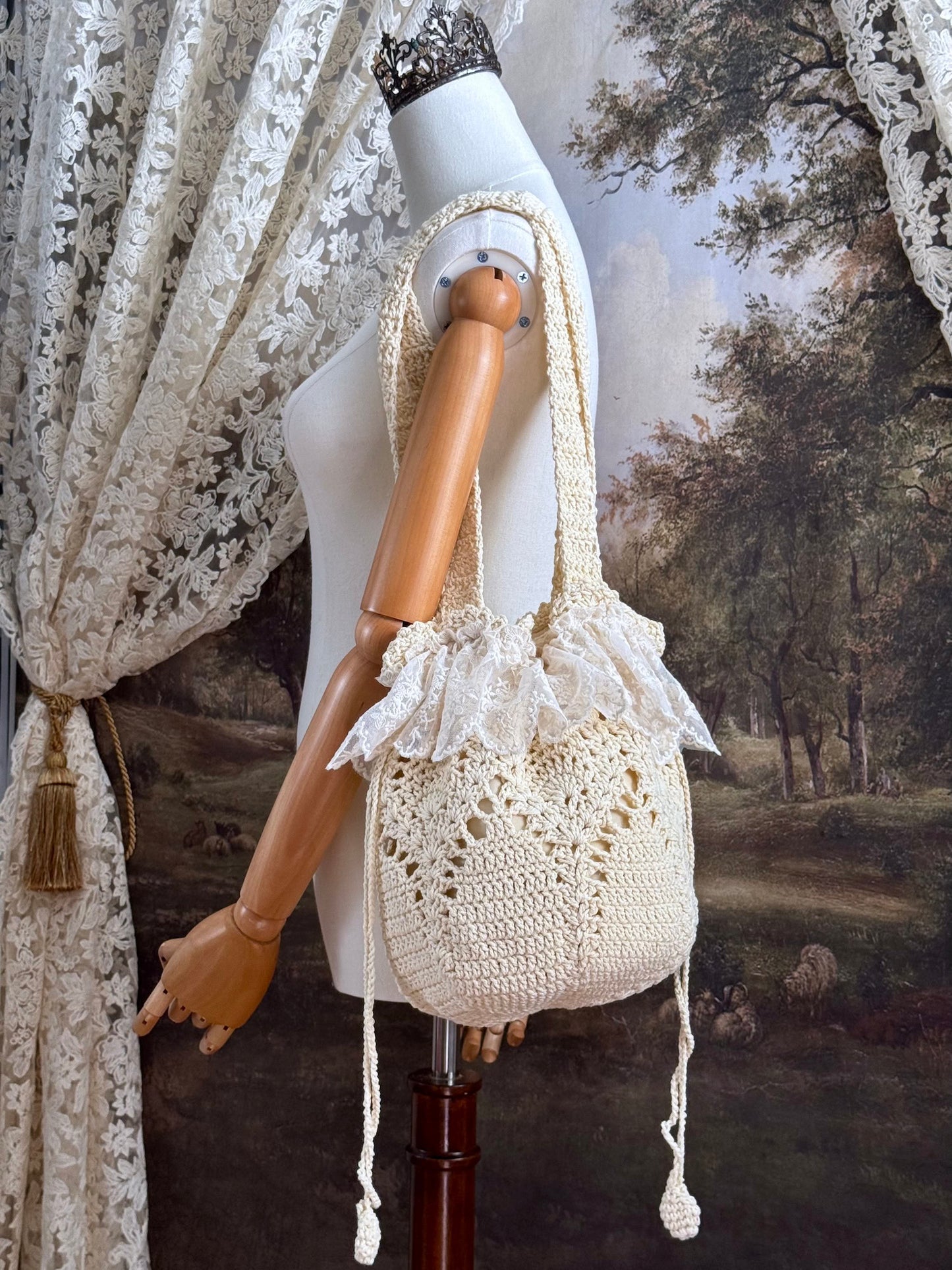 Vintage inspired crochet bucket shoulder bag with lace trim and drawstring closure, pictured on a mannequin in front of a historical painting backdrop.