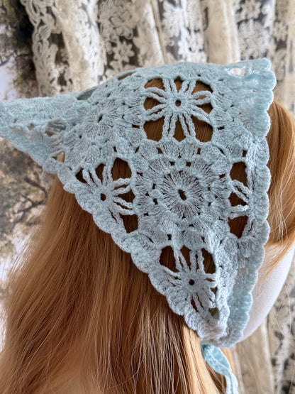 A vintage inspired light blue crochet babushka triangle headdress on a mannequin head in front of lace curtains.