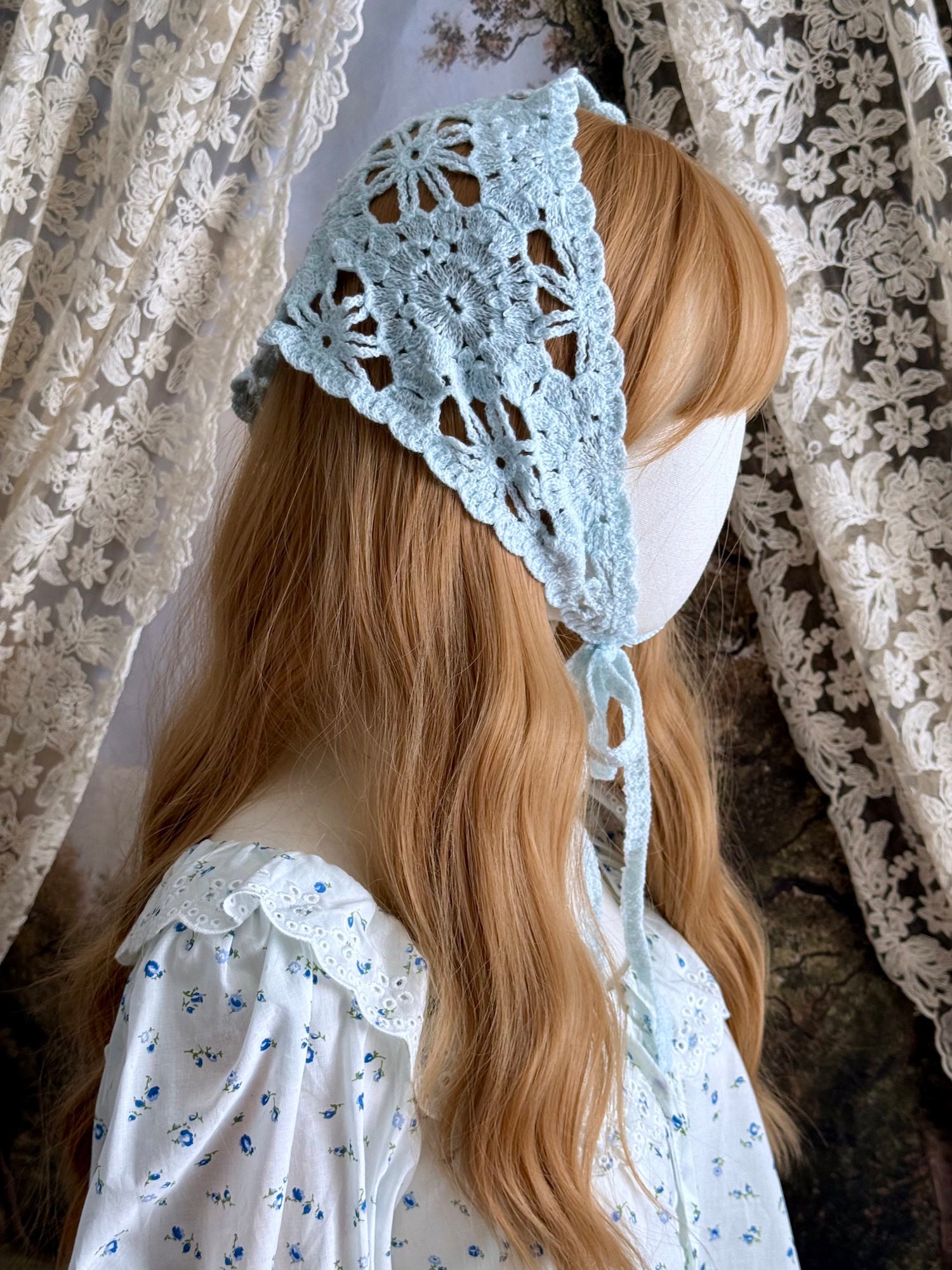 A vintage inspired light blue crochet babushka triangle headdress on a mannequin head in front of lace curtains.
