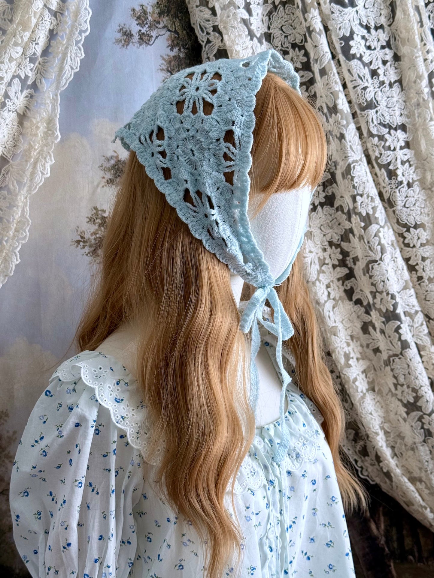 A vintage inspired light blue crochet babushka triangle headdress on a mannequin head in front of lace curtains.