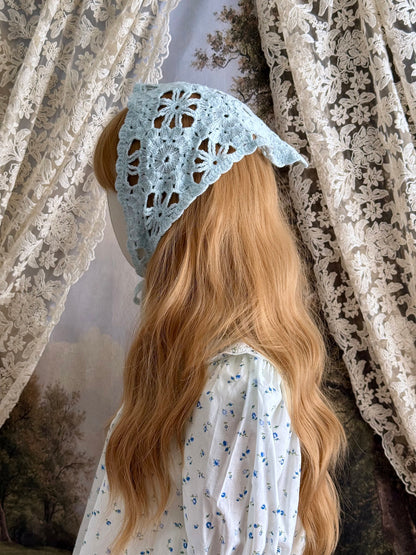 A vintage inspired light blue crochet babushka triangle headdress on a mannequin head in front of lace curtains.