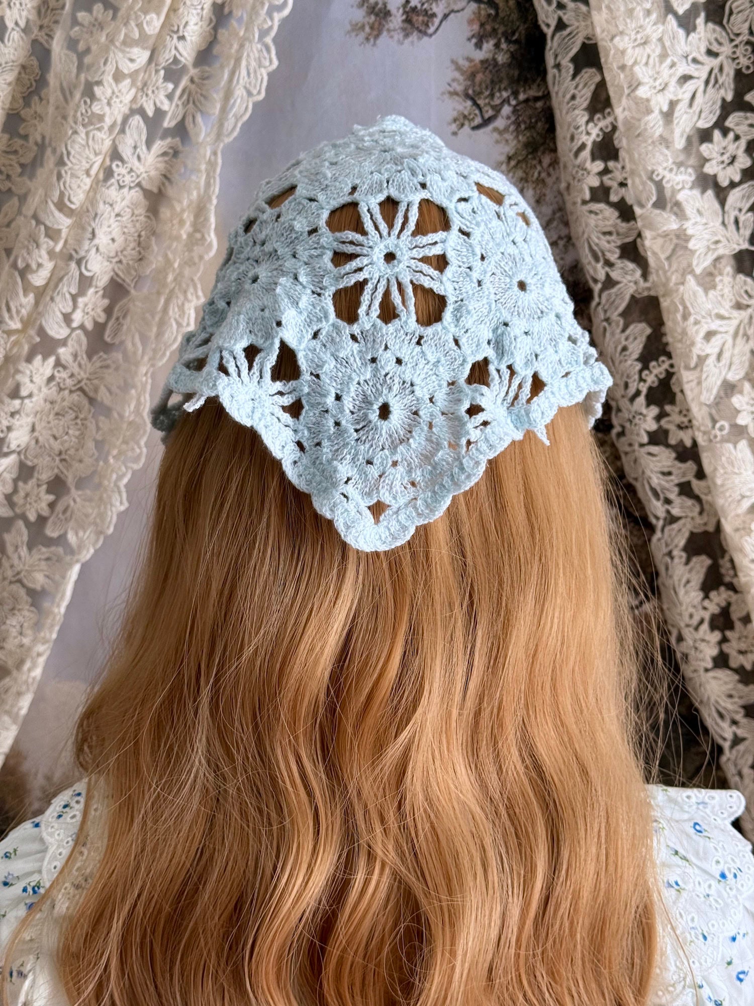 A vintage inspired light blue crochet babushka triangle headdress on a mannequin head in front of lace curtains.