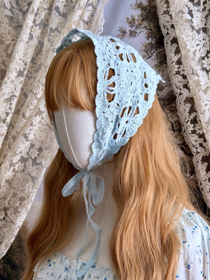 A vintage inspired light blue crochet babushka triangle headdress on a mannequin head in front of lace curtains.
