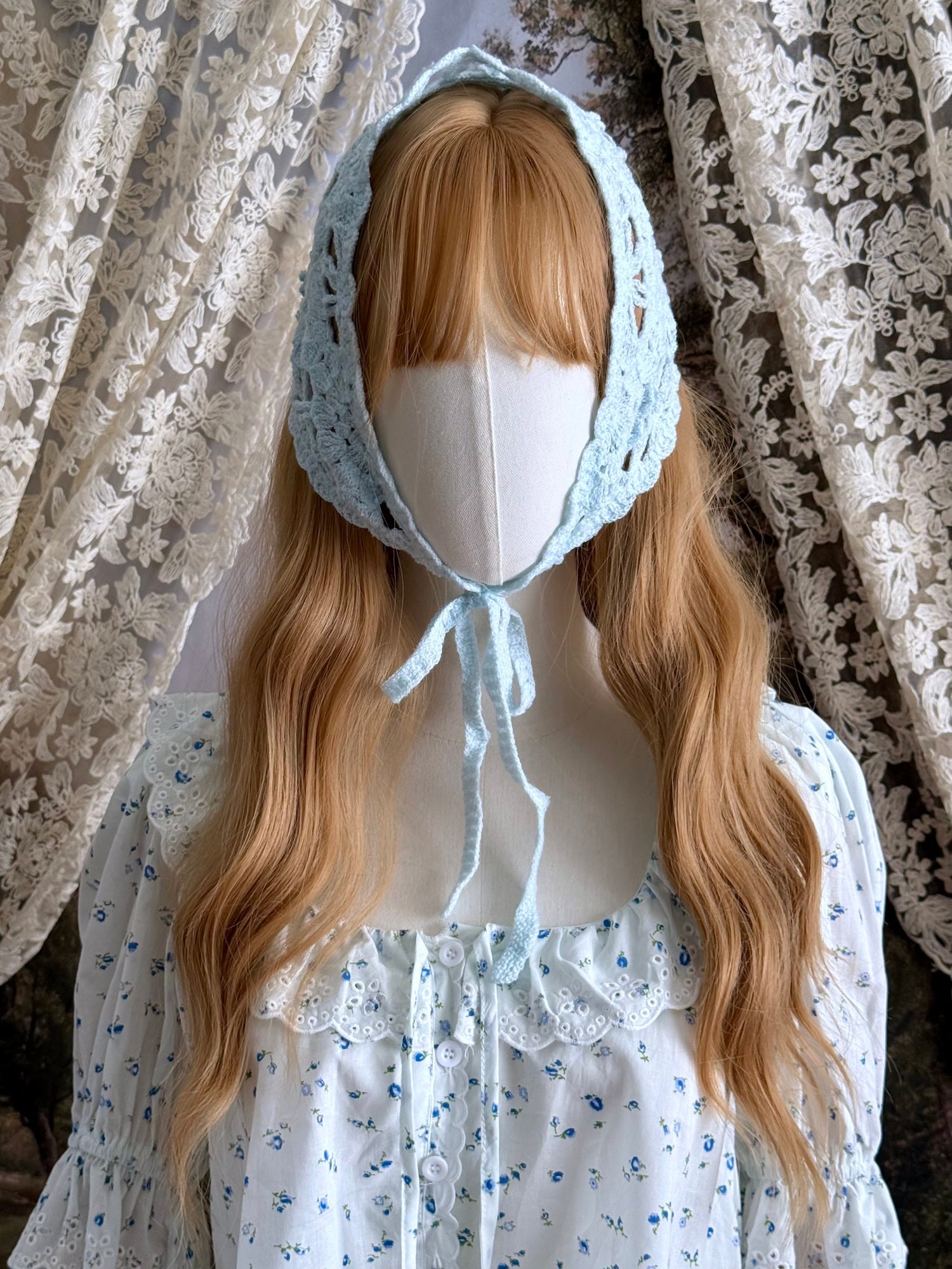 A vintage inspired light blue crochet babushka triangle headdress on a mannequin head in front of lace curtains.