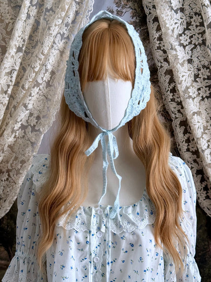 A vintage inspired light blue crochet babushka triangle headdress on a mannequin head in front of lace curtains.
