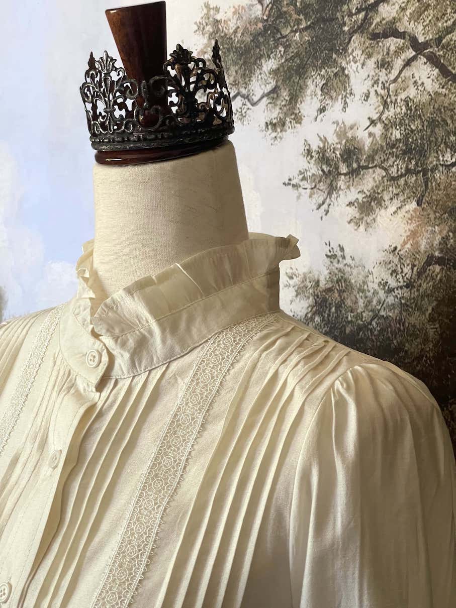 A historically inspired victorian and edwardian era style ivory colored blouse with pintucks, a high neck collar, ruffle hem sleeves, and crochet lace.