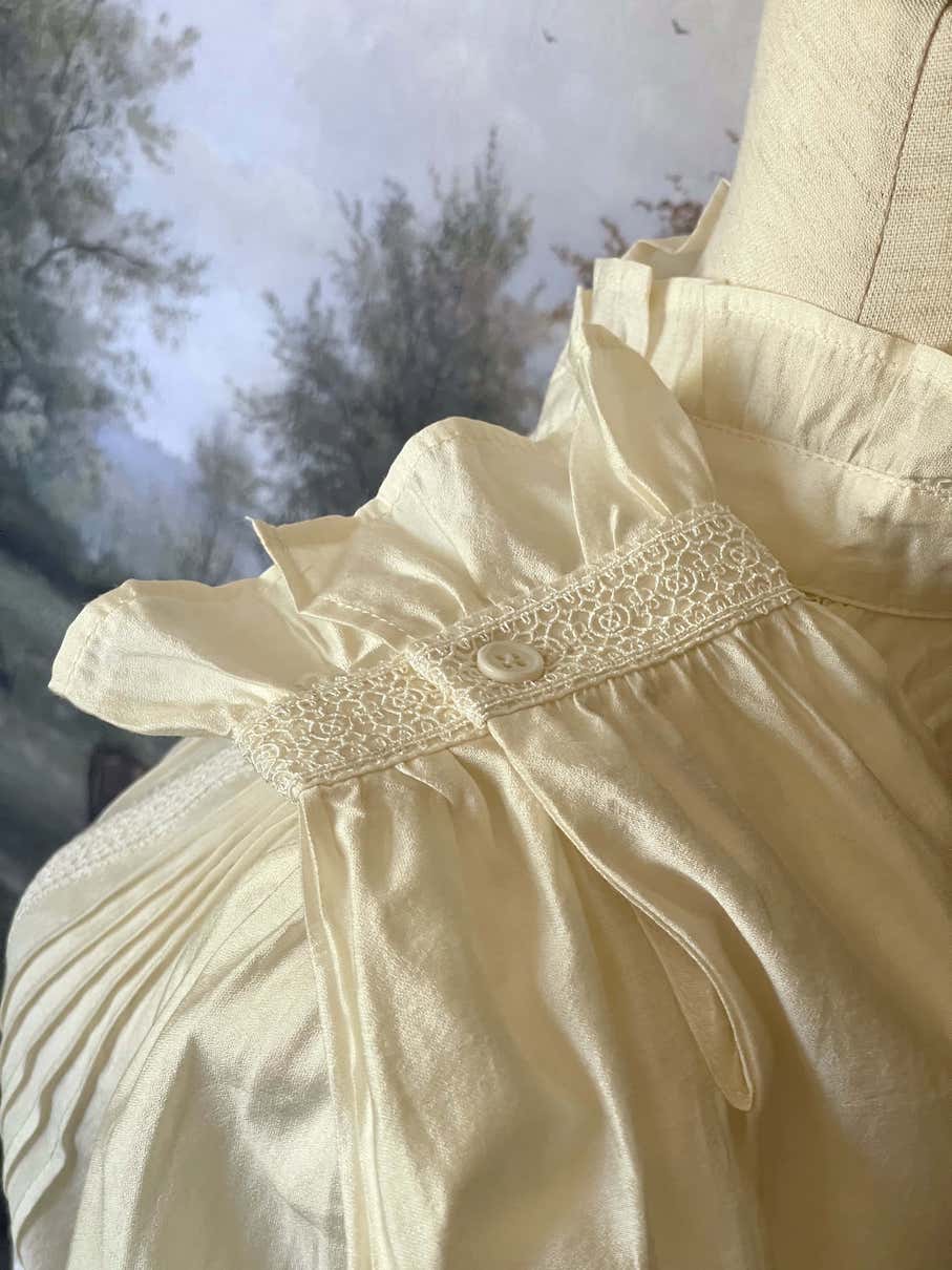 A historically inspired victorian and edwardian era style ivory colored blouse with pintucks, a high neck collar, ruffle hem sleeves, and crochet lace.
