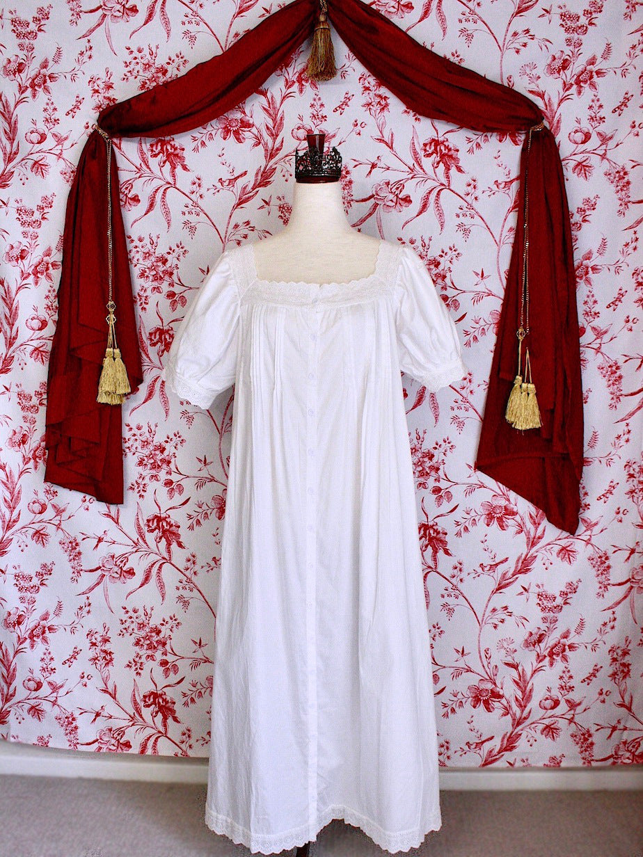A historically inspired regency style short sleeve chemise dress with pin-tuck pleats and lace trim in front of a toile backdrop.