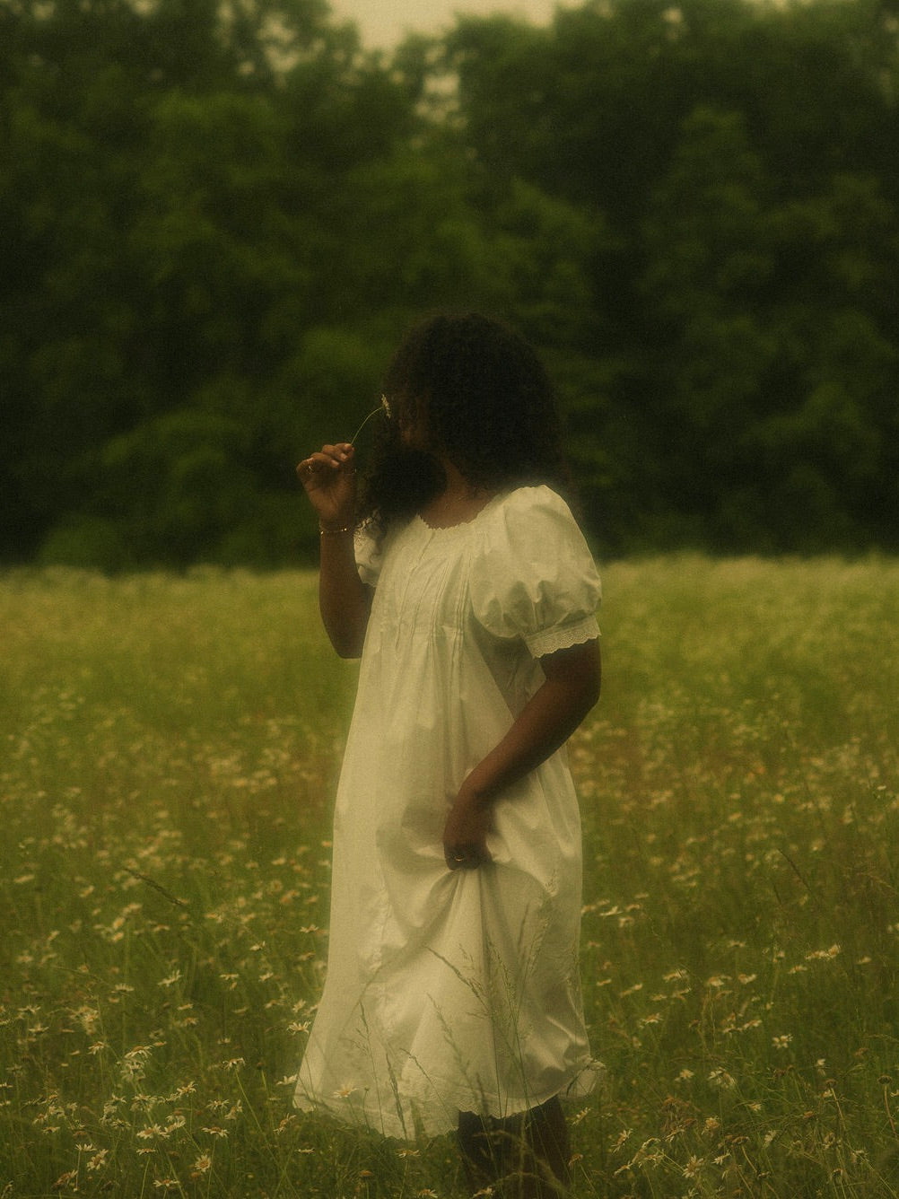 A woman in a flower field wearing a vintage style historically-inspired white short-sleeve chemise dress with lace trim and pleated details, holding a small flower.