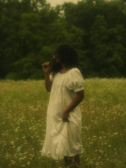 A woman in a flower field wearing a vintage style historically-inspired white short-sleeve chemise dress with lace trim and pleated details, holding a small flower.