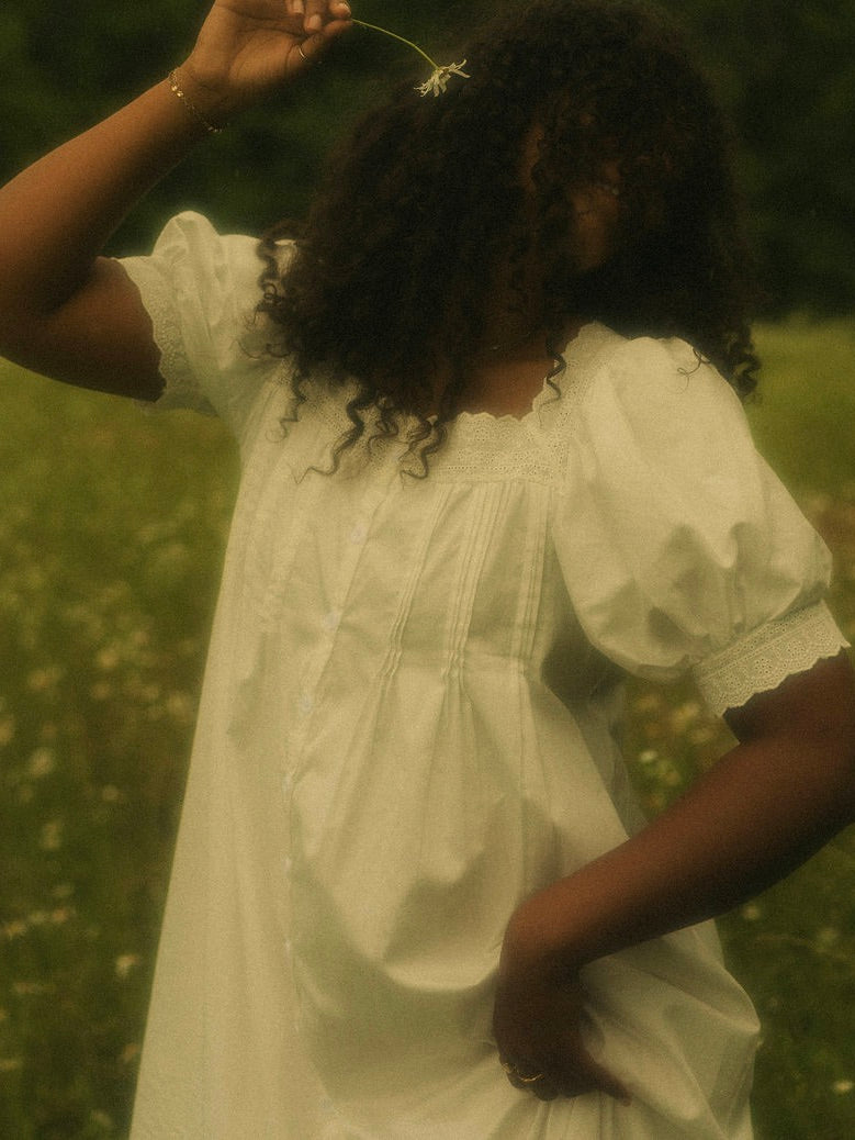 A woman in a flower field wearing a vintage style historically-inspired white short-sleeve chemise dress with lace trim and pleated details, holding a small white flower.