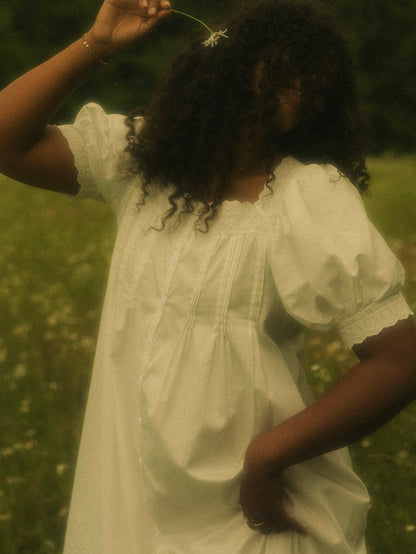 A woman in a flower field wearing a vintage style historically-inspired white short-sleeve chemise dress with lace trim and pleated details, holding a small white flower.