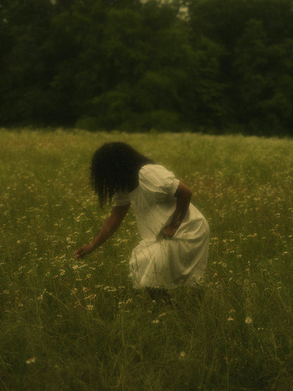 A woman in a flower field wearing a vintage style historically-inspired white short-sleeve chemise dress with lace trim, puff sleeves, and pleated details.