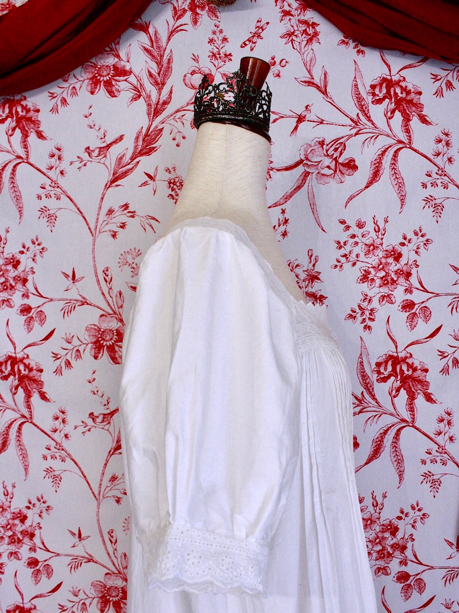 A historically inspired regency style short sleeve chemise dress with pin-tuck pleats and lace trim in front of a toile backdrop.