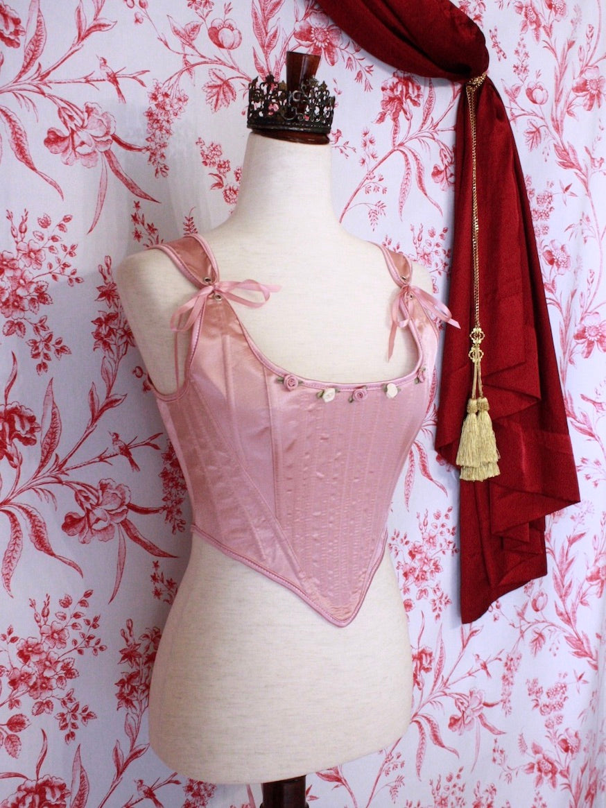 A pair of Balletcore Renaissance, Baroque, Rococo, Regency, Victorian Era Corset Stays in Ballet Pastel Pink with Rosette Appliques in front of a toile backdrop.