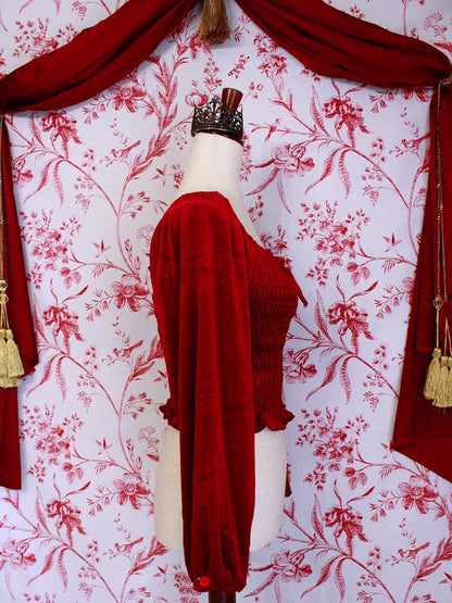 A Historically Inspired Renaissance to Victorian Era  Red Velvet Smocked Blouse with Rosette Bow Applique in front of a toile backdrop.