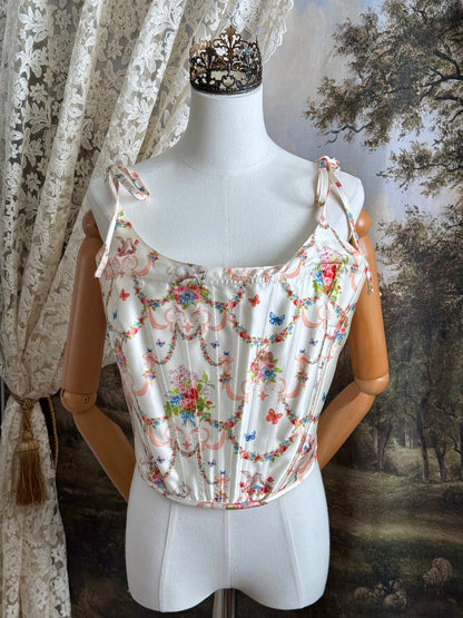 A historically-inspired 1700s rococo era corset top in an ivory floral printed satin is pictured on a mannequin in front of an art history backdrop.