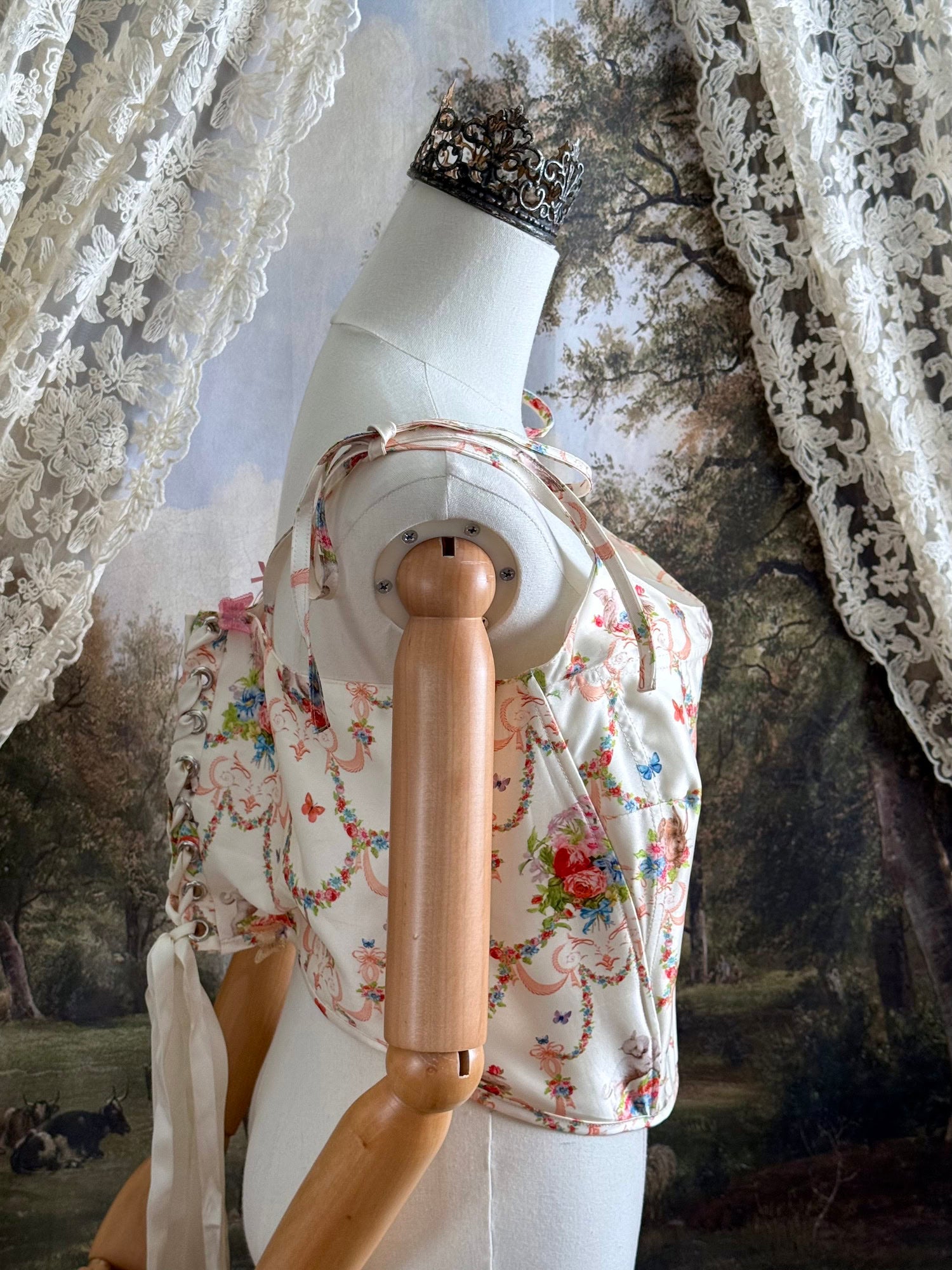 A historically-inspired 1700s rococo era corset top in an ivory floral printed satin is pictured on a mannequin in front of an art history backdrop.