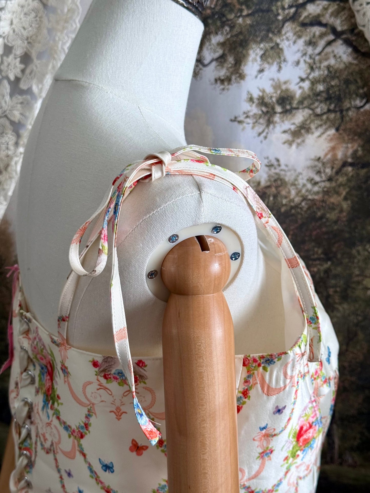 A historically-inspired 1700s rococo era corset top in an ivory floral printed satin is pictured on a mannequin in front of an art history backdrop.