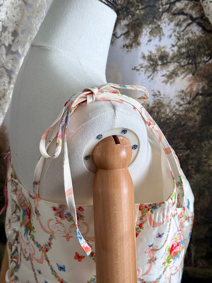A historically-inspired 1700s rococo era corset top in an ivory floral printed satin is pictured on a mannequin in front of an art history backdrop.
