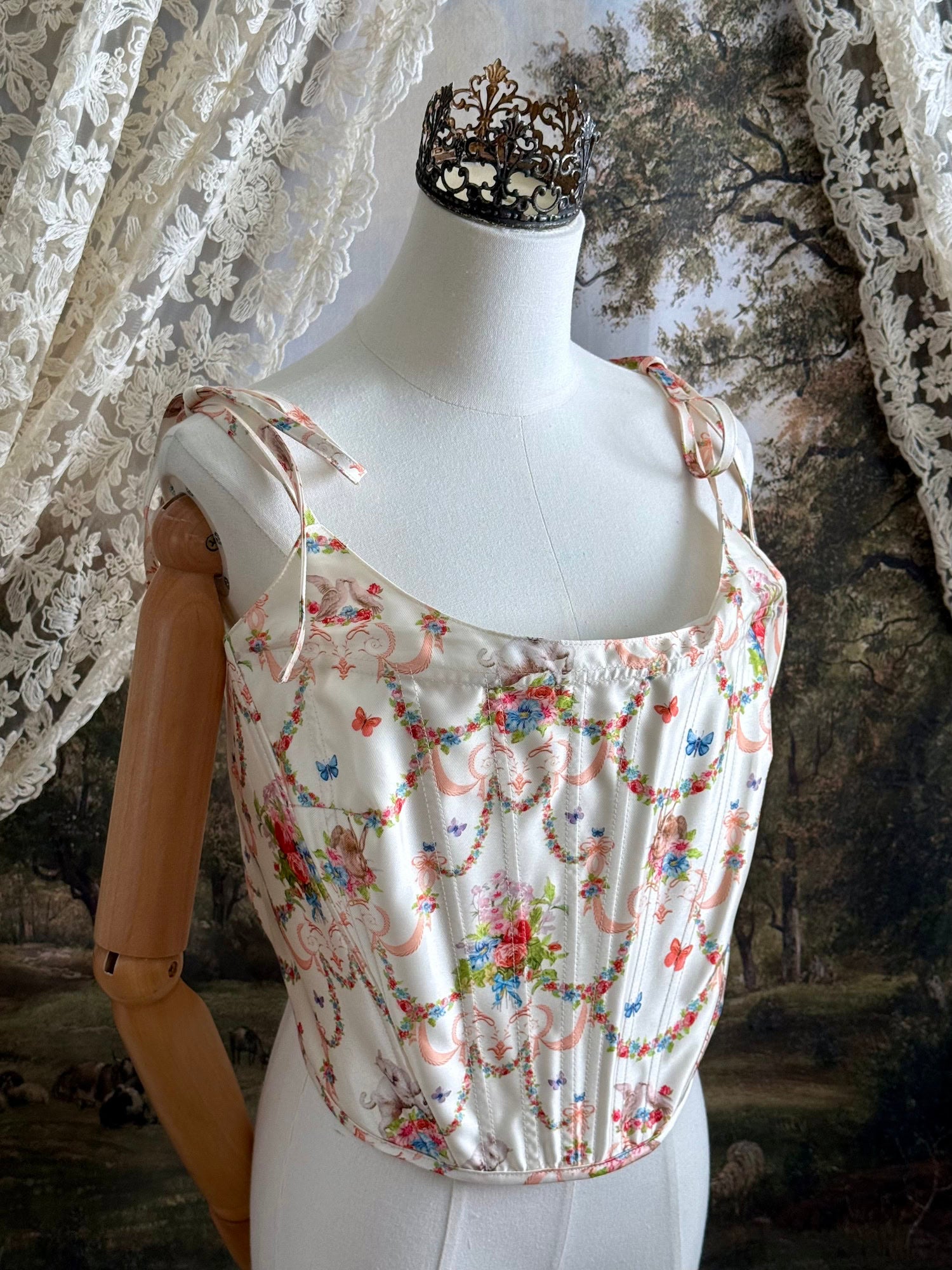 A historically-inspired 1700s rococo era corset top in an ivory floral printed satin is pictured on a mannequin in front of an art history backdrop.