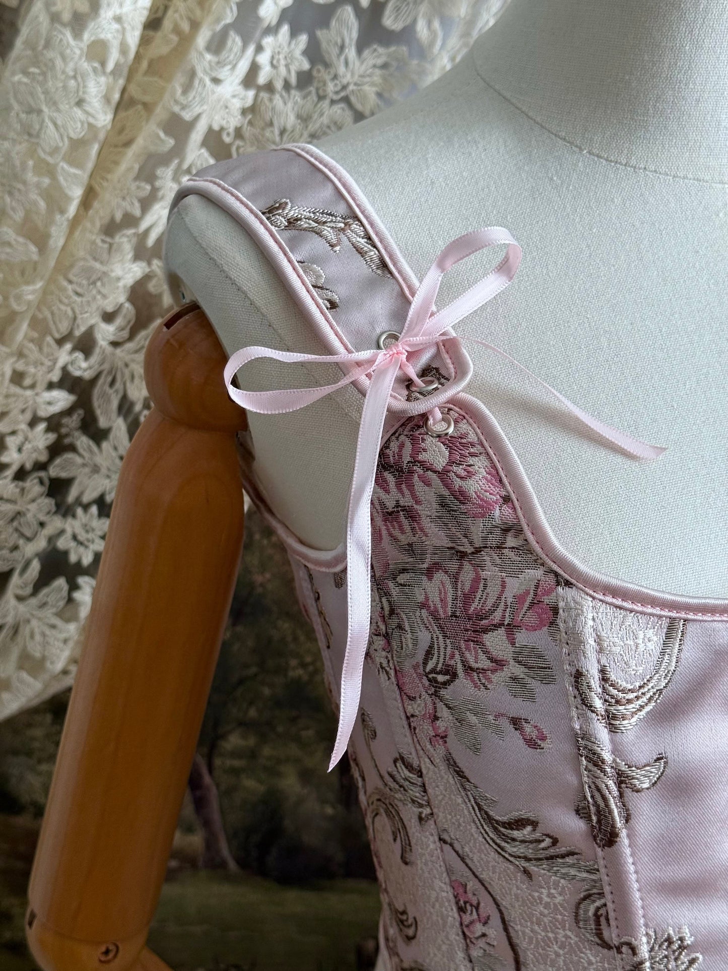 Historical fashion inspired baroque to rococo era front-laced corset stays top in pastel pink floral jacquard fabric, pictured on a mannequin. Detail of ribbons.