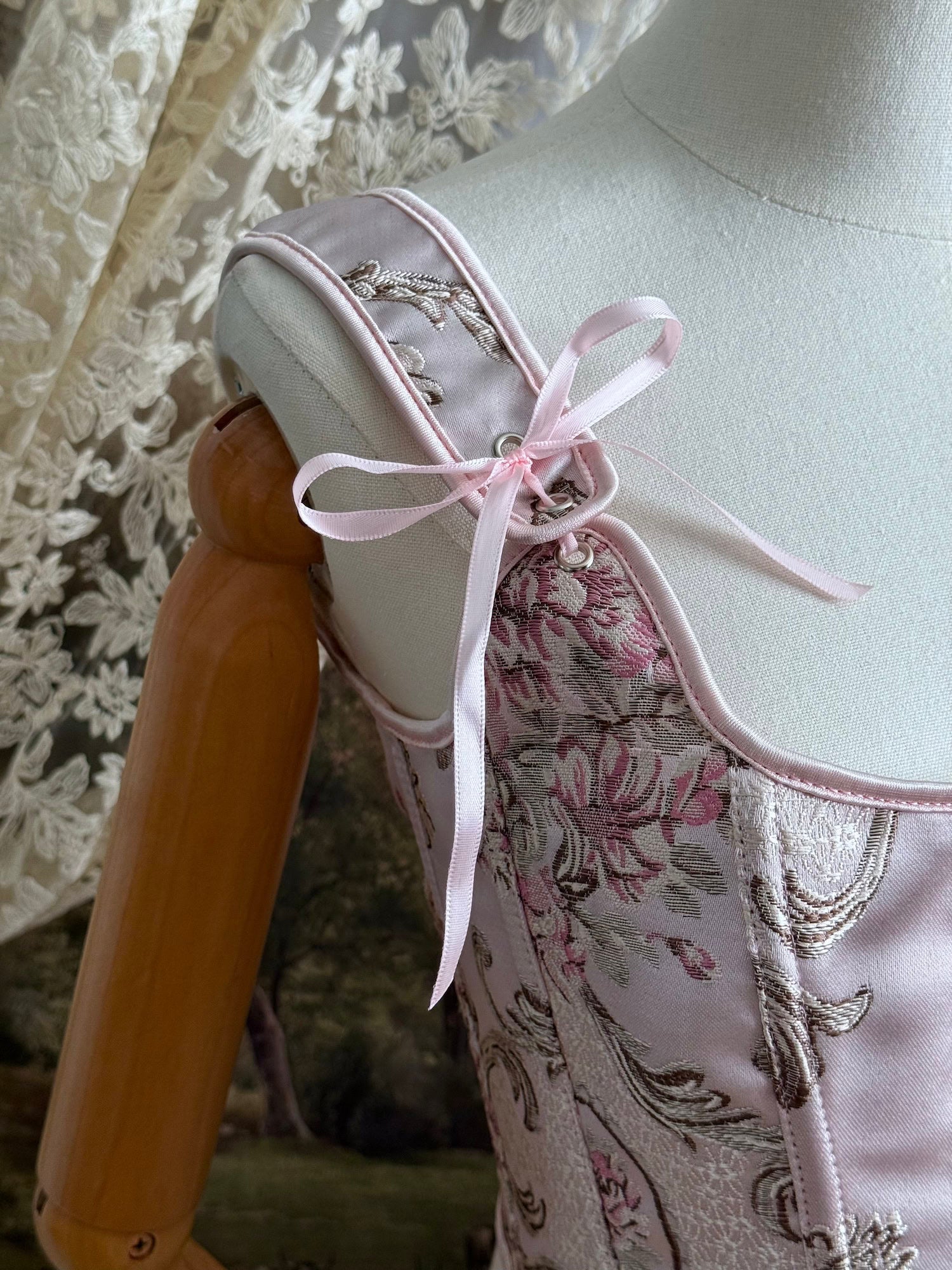 Historical fashion inspired baroque to rococo era front-laced corset stays top in pastel pink floral jacquard fabric, pictured on a mannequin. Detail of ribbons.