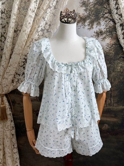A set of vintage inspired white and blue floral print cotton bloomer shorts and matching blouse with scalloped lace trim and bow-tied waist drawstring, pictured on a mannequin.