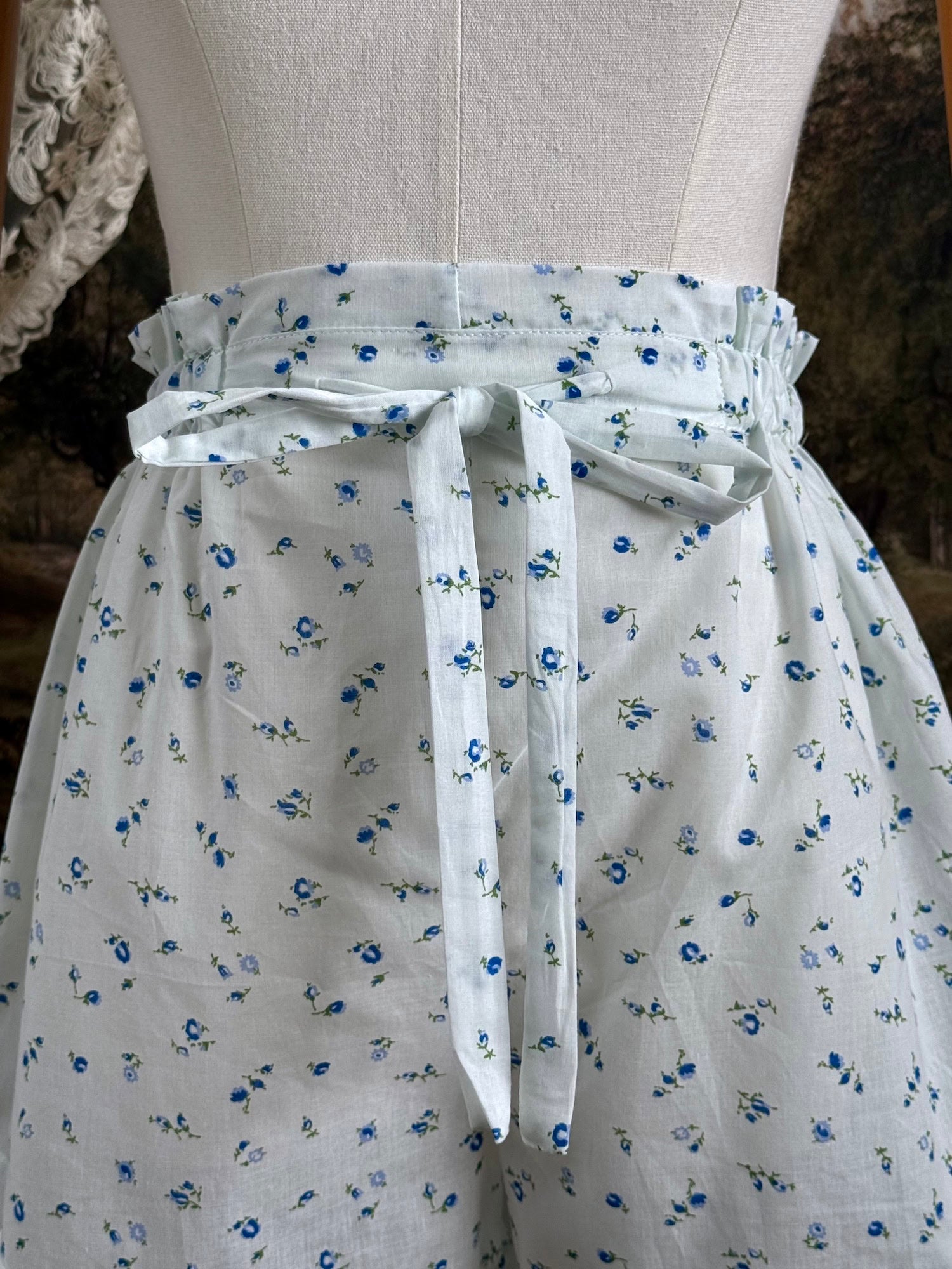 A pair of vintage inspired white and blue floral print cotton bloomer shorts with scalloped lace trim and bow-tied waist drawstring, pictured on a mannequin.