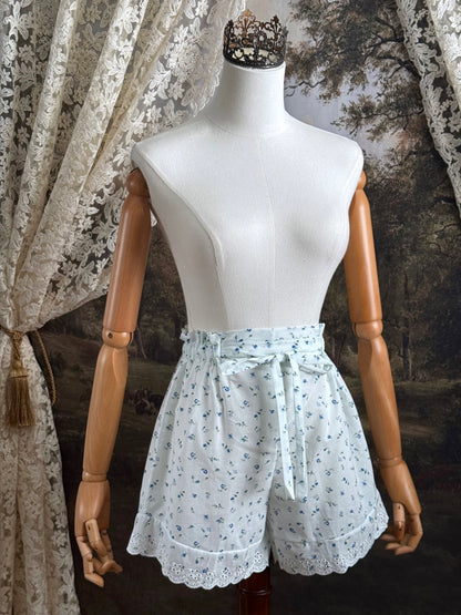 A pair of vintage inspired white and blue floral print cotton bloomer shorts with scalloped lace trim and bow-tied waist drawstring, pictured on a mannequin.