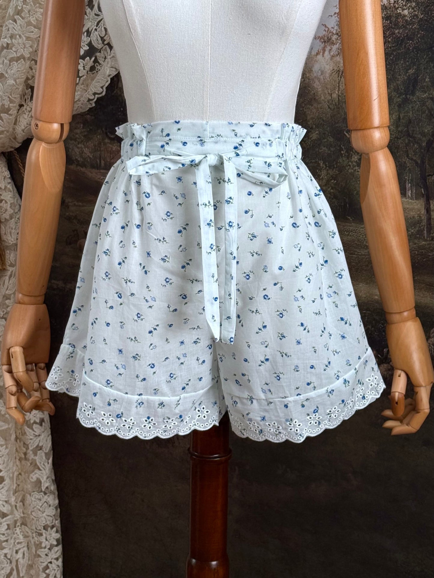 A pair of vintage inspired white and blue floral print cotton bloomer shorts with scalloped lace trim and bow-tied waist drawstring, pictured on a mannequin.